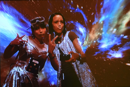 Amber and a friend in cheap silver costumes, making silly duck faces and throwing peace signs in front of a picture of space. A filter makes their colouring look alien.